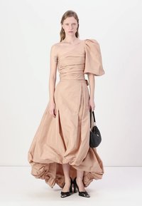 Beige mekko, jossa on epäsymmetrinen leikkaus, muotoiltu yläosa, yksi puffihihna ja rypistetty hame. Asusteena musta käsilaukku ja korkokengät.