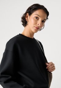 Felpa nera con colletto rotondo e vestibilità morbida; texture liscia. Il modello tiene una borsa a spalla con una tonalità attenuata, posizionata su uno sfondo neutro.