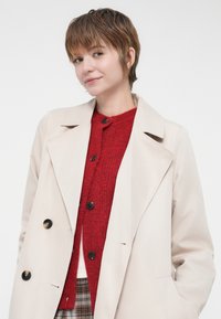 Κρεμ παλτό με μεγάλα μαύρα κουμπιά, φορεμένο πάνω από μια κόκκινη πλεχτή μπλούζα. Λείο ύφασμα, μαζεμένο γιακά και κομμένο ράψιμο.