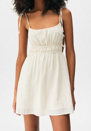 Vestito beige chiaro con spalline sottili regolabili, vita elastica, tessuto fluido e ritagli laterali per un dettaglio in più. Materiale morbido e testurizzato.