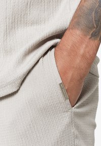 Δεξί χέρι με τατουάζ που εισάγεται σε τσέπη από υφή ανοιχτού μπεζ υφάσματος με μεταλλική ετικέτα που γράφει "CLOSURE LDN."