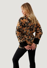 Top envolvente marrón con estampado floral y acentos negros, con mangas largas y un detalle de cinturón, combinado con jeans ajustados negros.