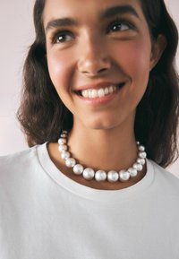 Masivna srebrna biserna ogrlica z različno velikostjo, gladka tekstura, v kombinaciji s preprosto belo majico in nevtralno ozadje.