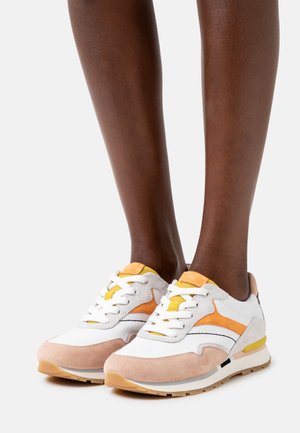 Pari valkoisia, beigen ja oranssin sävyisiä tennareita, joita käytetään tummalla iholla olevilla jaloilla, seisomassa yksinkertaista vaaleaa taustaa vasten.