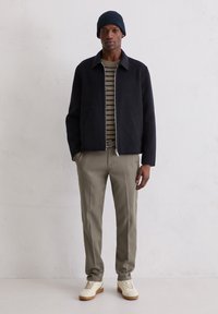 Granatowa kurtka zapinana na zamek na swetrze w paski, zestawiona z oliwkowozielonymi spodniami i białymi tenisówkami. Model nosi także ciemną dzianinową czapkę.