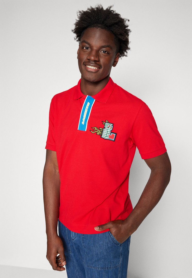 Polo rouge avec un col bleu présentant un texte de logo. L'avant arbore un design brodé avec des flammes et un graphique. Tissu en coton doux.