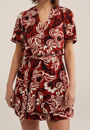 Femme portant une robe à manches courtes avec col en V, motif floral rouge, noir et blanc et ceinture nouée à la taille, debout devant un fond beige.