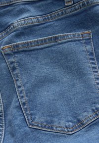 Blugii din denim au o țesătură albastră cu o textură ușor estompată, cusături vizibile și un buzunar posterior cusut cu un fir de culoare contrastantă.