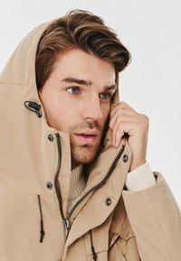 Beżowy wodoodporny parka z zamkiem błyskawicznym i zapięciem na troczek, z wysokim kołnierzem i kapturem. Posiada czarne elementy metalowe i strukturalne wnętrze.