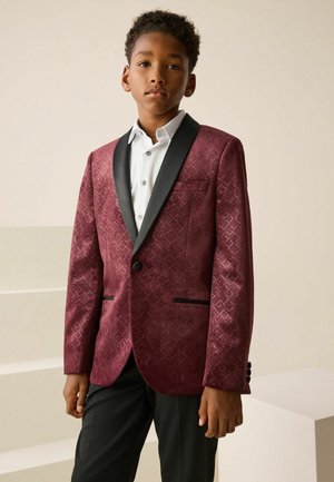 Dreng iført en tekstureret bordeaux blazer med sorte revers, hvid skjorte og sorte bukser står foran en neutral baggrund.