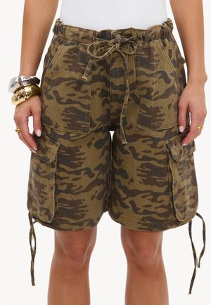 Short cargo camouflage ample avec poches avant, taille à cordon et poches latérales à rabat, porté par une personne avec des bracelets et des bagues.