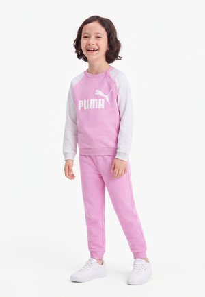Enfant aux cheveux courts et foncés souriant, portant un sweat-shirt Puma rose et gris, un pantalon rose assorti et des baskets blanches, debout sur fond blanc.
