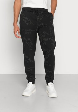 Mann trägt schwarze Calvin Klein Jeans Jogginghose mit Kordelzug in der Taille und weiße Sneakers, steht auf grauem Boden vor einer weißen Wand.