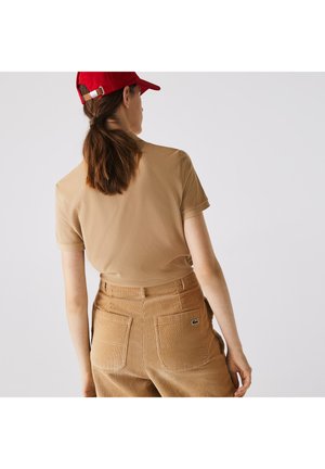 Chemise à manches courtes marron côtelée assortie à un pantalon en velours côtelé beige texturé. Casquette rouge avec sangle réglable à l'arrière. Logo Lacoste sur le pantalon.