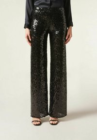 Pantalons larges en sequins noirs avec taille haute. Comprend des sequins brillants et réfléchissants ainsi qu'une texture lisse. Associés à des sandales noires à brides.