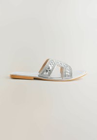 Sandale plate argentée à bride glissée, avec bout ouvert, sangle tissée décorative et semelle beige sur fond clair uni.