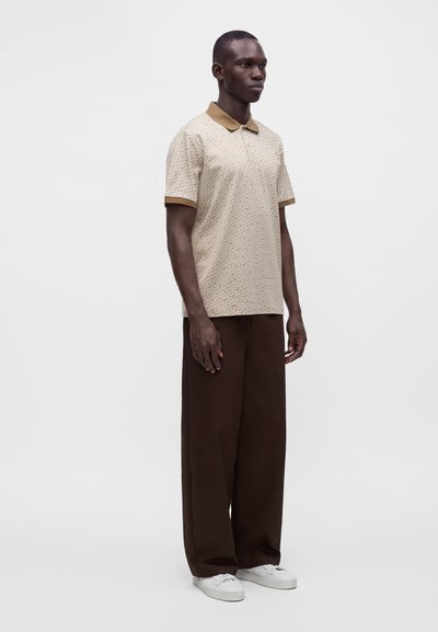 Homme debout portant un polo beige à manches courtes à motif, un pantalon large marron foncé et des baskets blanches sur un fond blanc uni.