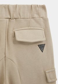 Beżowe dzianinowe spodnie z elastycznym pasem, z klapką kieszeni z boku, dodatkową klapką kieszeni w pobliżu dołu oraz czarną naszywką logo Guess.