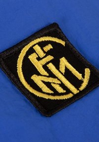 Textielpatch met een cirkelvormige zwarte achtergrond, versierd met gouden geborduurde letters "F" en "M" in een gestileerd lettertype, op een blauwe stofoppervlak.