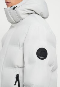 Person iført en lysegrå pufferjakke med høj krave, hætte, sort lynlås og sort cirkulært mærke på overarmen.