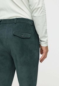 Jobb oldali nézet egy személyről, aki sötétzöld kordbársony nadrágot visel, gombos hátsó zsebbel és világos, hosszú ujjú felsővel egy sima háttér előtt.