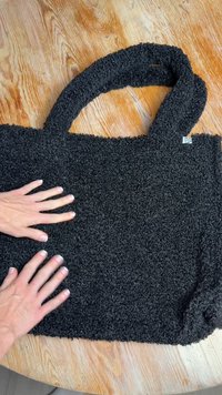 Mustast, tekstuuriga kangast ostukott, millel on kaks sangast, valmistatud pehmest kangast. Alumises paremas nurgas on silt. Kotti toetuvad käed.