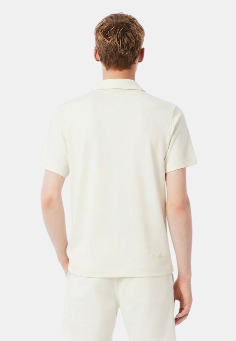 Man met kort haar, gekleed in een eenvoudige, off-white korte mouw shirt en bijpassende korte broek, staand met zijn rug naar de kijker op een witte achtergrond.