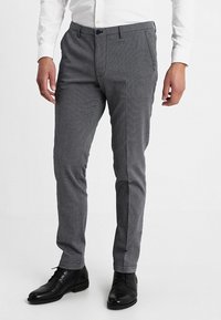 Šedé kalhoty s rybí kostí vzorem z tkané látky, s úzkým střihem, bočními kapsami a zapínáním na knoflíky, doplněné černými botami.