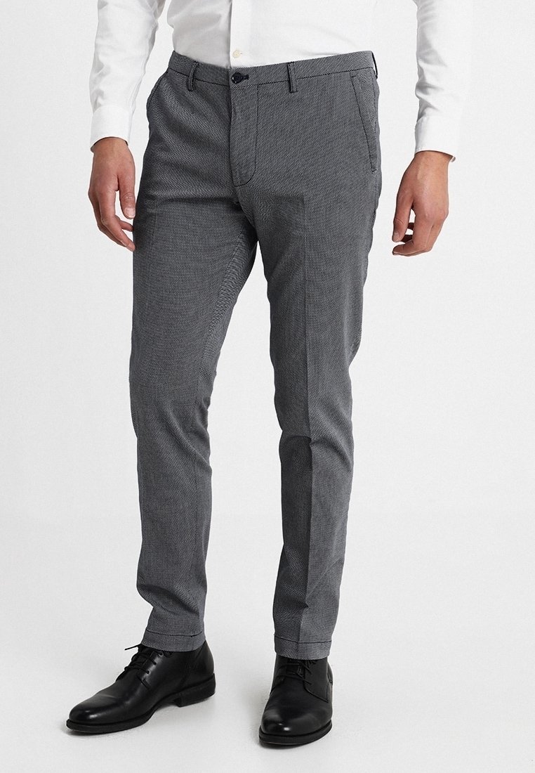 Šedé kalhoty s rybí kostí vzorem z tkané látky, s úzkým střihem, bočními kapsami a zapínáním na knoflíky, doplněné černými botami.
