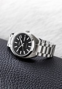 Αυτόματο ρολόι χειρός Timex Marlin ασημί, με μαύρο καντράν, λευκούς δείκτες ώρας και ημερομηνία ημέρας, τοποθετημένο σε υφή μαύρης επιφάνειας.