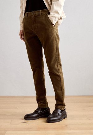 Homme portant un pantalon en velours côtelé marron et des mocassins en cuir brun foncé debout sur un sol en bois.