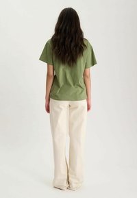 T-shirt vert olive à manches courtes en coton, associé à un pantalon large couleur crème en tissu lisse et texturé.