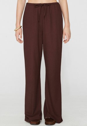 Pantalones marrones de pierna ancha con cintura con cordón, combinados con zapatos mocasines marrones y una pulsera de oro en la muñeca izquierda.