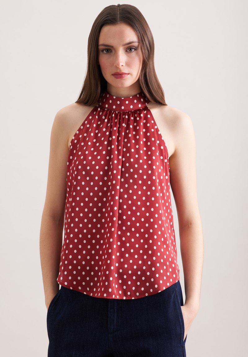 Femme portant un haut à bretelles rouge à pois blancs et un pantalon bleu marine, debout devant un fond clair uni.