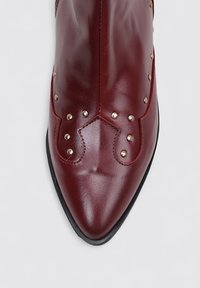 Bottine en cuir bordeaux avec un bout pointu, ornée de clous métalliques et d'une texture lisse, reposant sur une semelle en caoutchouc noir.