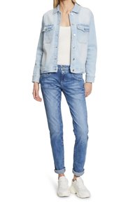 Chaqueta de mezclilla azul clara con dos bolsillos en el pecho y botones plateados, combinada con jeans azul claros y zapatillas blancas. Corte informal y mangas remangadas.