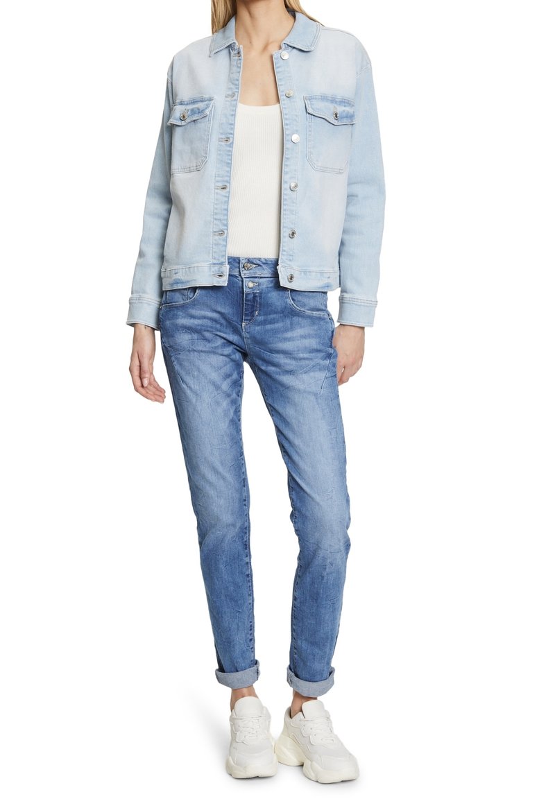 Chaqueta de mezclilla azul clara con dos bolsillos en el pecho y botones plateados, combinada con jeans azul claros y zapatillas blancas. Corte informal y mangas remangadas.