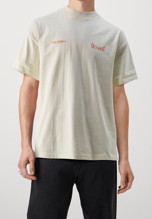 Person trägt ein cremefarbenes kurzärmeliges T-Shirt mit rotem Text "1980's AMERICA" und "OUT WEST" sowie schwarze Hose, Hände entspannt an den Seiten.