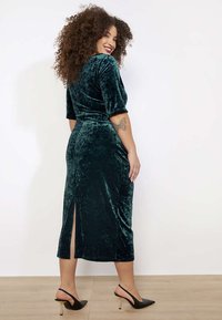 Zelené semišové midi šaty s krátkymi rukávmi a rozparkom na chrbte. Vyznačujú sa hladkou textúrou a priliehavým strihom. Kombinované s čiernymi lodičkami.