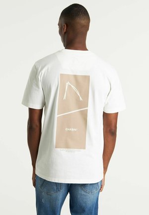 Mężczyzna noszący białą koszulkę z beżową prostokątną grafiką i napisem "CHASIN'" na plecach, w połączeniu z niebieskimi dżinsami.