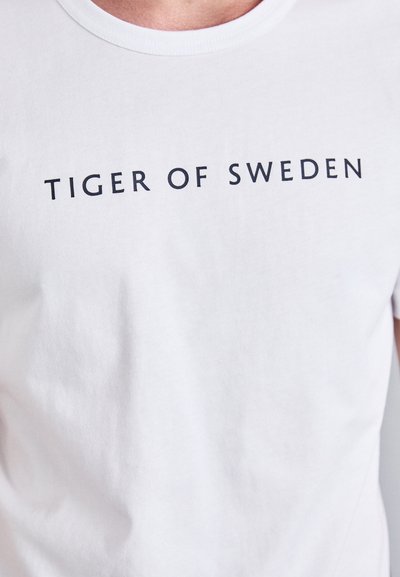 Hvit bomull t-skjorte med fet svart tekst som sier "TIGER OF SWEDEN" over brystet. Klassisk rund hals-design, glatt tekstur.