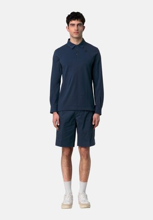 Uomo in piedi rivolto in avanti che indossa una polo blu navy a maniche lunghe, pantaloncini blu navy fino al ginocchio, calzini bianchi e scarpe da ginnastica bianche.