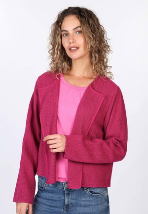 Fuchsia knitted jacket with an open front, side pockets, and wide sleeves, layered over a pink blouse, paired with blue jeans.