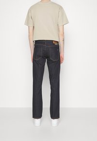 Personne portant un t-shirt beige, un jean bleu foncé avec des poches arrière, et des baskets blanches, se tenant face à un mur blanc les bras détendus.