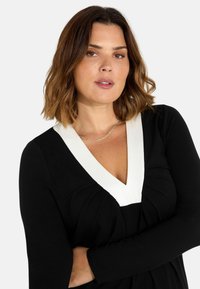 Musta mekko pitkillä hihoilla, V-pääntie, jossa on valkoinen yksityiskohta, rypyttämällä kaarrokeosa, sileä pinta. Minimalistinen muotoilu.