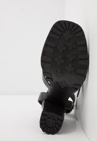 Sandale noire à talon reposant sur le côté, montrant une semelle robuste avec des motifs en forme de croix et de barreau sur un fond blanc d’angle.