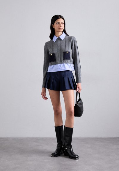 Suéter corto de canalé gris con bolsillos oscuros, superpuesto a una camisa azul clara. Una falda de tablas navy y botas negras hasta la rodilla completan el look.