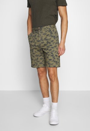 Mann trägt olivgrüne gemusterte Shorts, dunkelgrünes T-Shirt, weiße Socken und weiße Sneakers, steht mit den Händen in den Taschen.