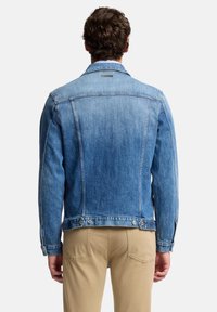 Mężczyzna w jasnoniebieskiej kurtce dżinsowej i beżowych spodniach, widziany od tyłu na jednolitym tle.