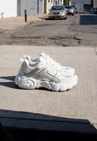 Een paar witte sportschoenen met dikke zolen geplaatst op een betonnen stoep vlakbij een straat met geparkeerde auto's in daglicht.